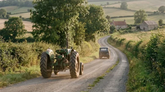 Vue arrière d'un tracteur sur une petite route de campagne bordée d'arbres avec une voiture patientant derrière